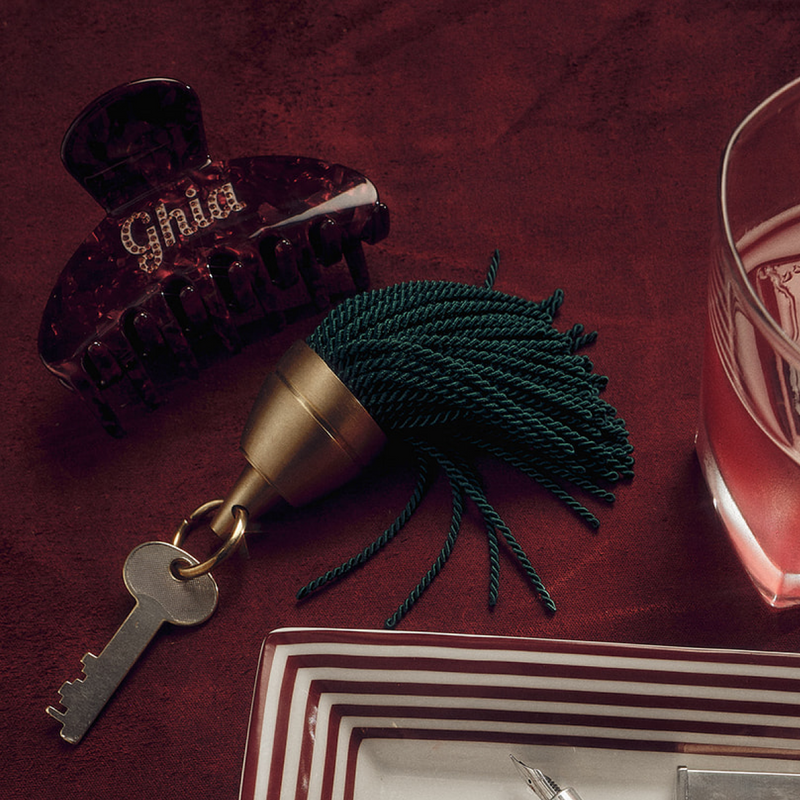 Burgundy Ghia claw clip on a table beside a brass bell with green tassel, a key, and a glass of red aperitif