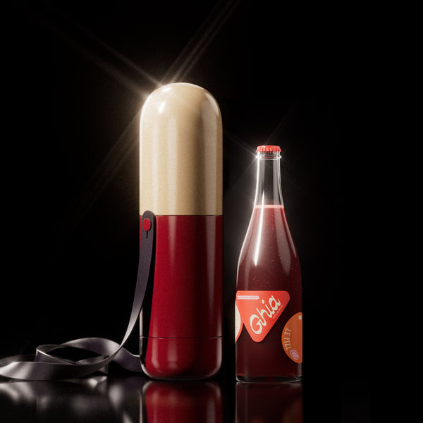 Cream and red pill-shaped Ghia carrier with strap beside a bottle of Ghia Le Fizz on a black background