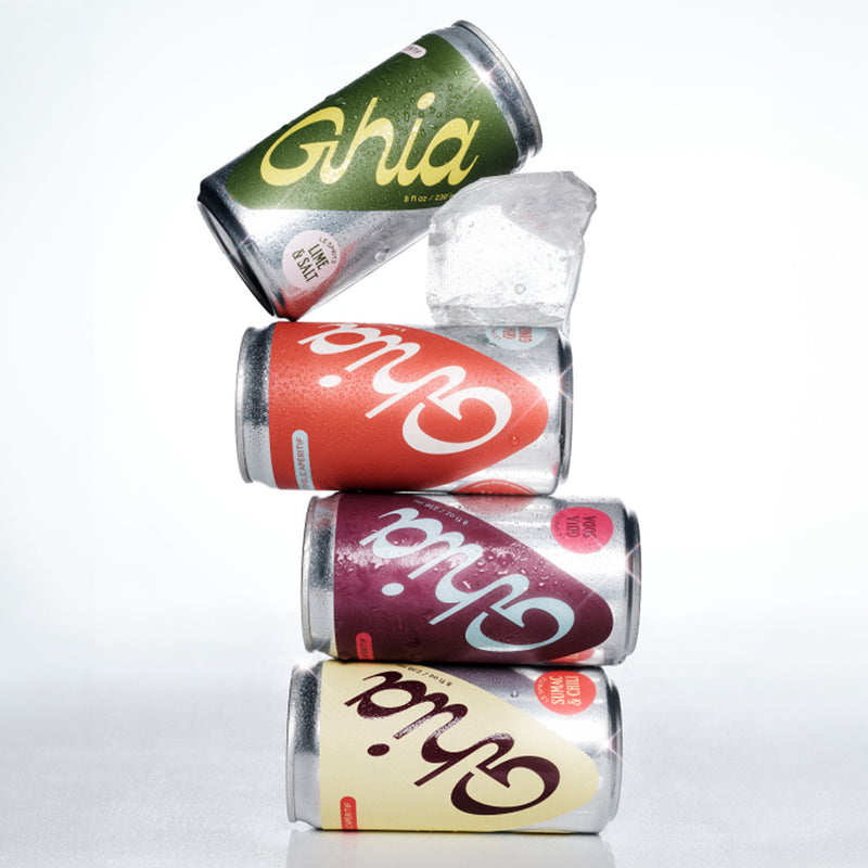 A stack of four cans of Ghia non-alcoholic aperitif in different flavors: lime, spritz, berry, and lemon, with droplets of condensation on the cans and an ice cube in the background.
