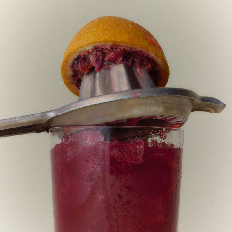 Blood orange half on a juicer pressed over a glass of deep red Ghia spritz